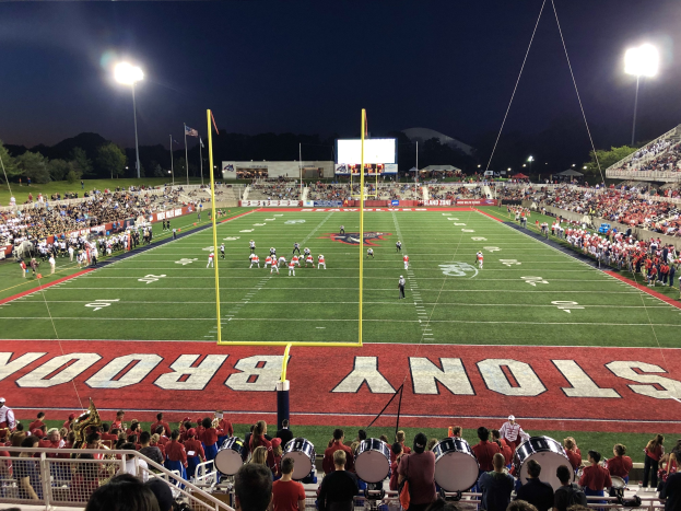 Ein nächtliches Fußballspiel in einem Stadion mit Spielern auf dem Feld, Zuschauern, Musikern, einem Metallzaun, Laternen, Fahnen, einem Anzeigebildschirm, Bäumen, Hügeln und einem sternenklaren Himmel.