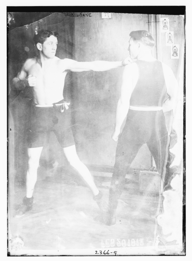 Schwarzes und weißes Foto von zwei Boxern in einem Ring mit Plakaten an der Wand dahinter und Text unten.