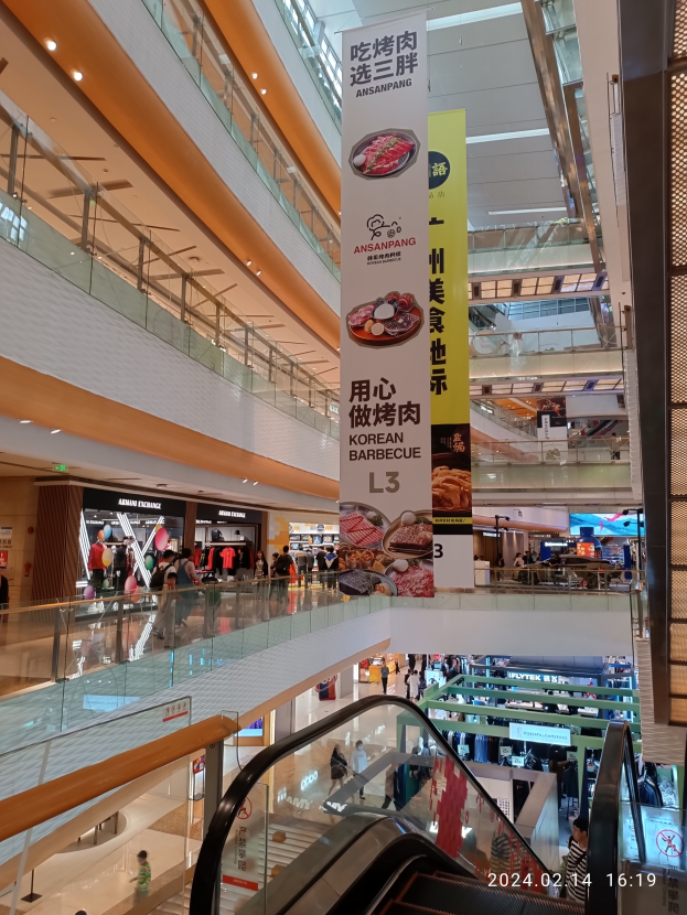 Einblicke in ein Einkaufszentrum mit einer Rolltreppe in der Mitte, Menschen mit Handtaschen, von der Decke hängende Banner und Geschäfte im Hintergrund.