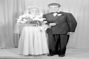 Ein Mann und eine Frau stehen nebeneinander auf einer Bühne, lächeln und halten Blumen in der Hand, mit einem Vorhang im Hintergrund.