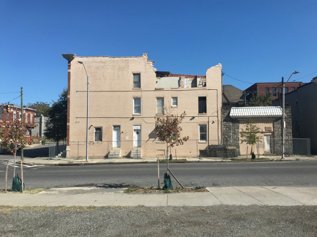 Ein altes, baufälliges Gebäude an einer Straßenecke mit zerbrochenen Fenstern, bröckelnden Wänden, umgeben von Straßenlaternen, Strommasten, Kabeln, Fahrzeugen, Bäumen und einem bewölkten Himmel.