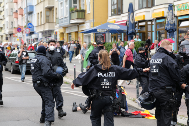 Eine Gruppe von Polizisten geht eine Straße entlang neben einer Menge mit Schirmen, Helmen und Handschuhen, mit Fahrzeugen, Schildern, Pflanzen und Gebäuden im Hintergrund.