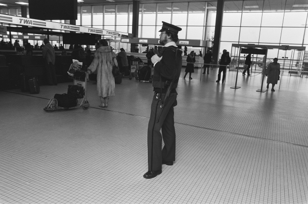 Polizist in Uniform mit Waffe in einem Flughafen-Terminal umgeben von Menschen und Gepäck.