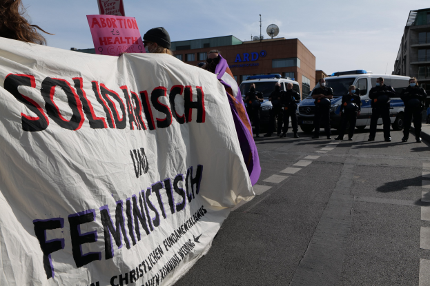 Gruppe von Menschen marschiert auf einer Straße und hält ein 'Solidarität und Feminismus'-Schild, mit geparkten Fahrzeugen und Gebäuden im Hintergrund unter einem klaren blauen Himmel.