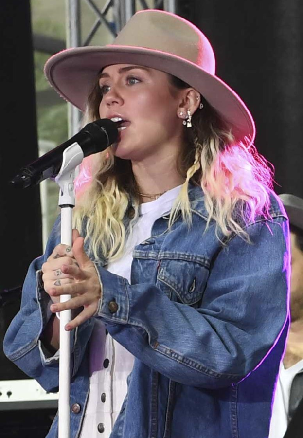 Miley Cyrus performs bei der CMA Music Festival in Nashville, Tennessee, singt in ein Mikrofon mit Ständer und trägt eine Mütze, während eine Person mit einer Kappe neben ihr steht und musikalische Instrumente im Hintergrund zu sehen sind.