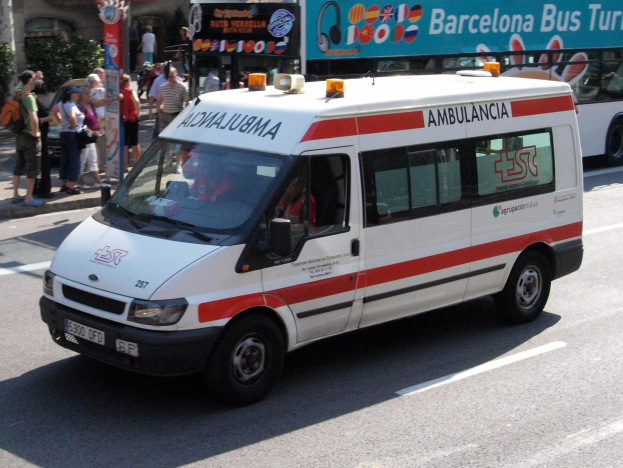 Rettungswagen fährt neben einem Bus mit Menschen auf dem Gehweg, Gebäude und Bäume im Hintergrund unter einem klaren blauen Himmel.