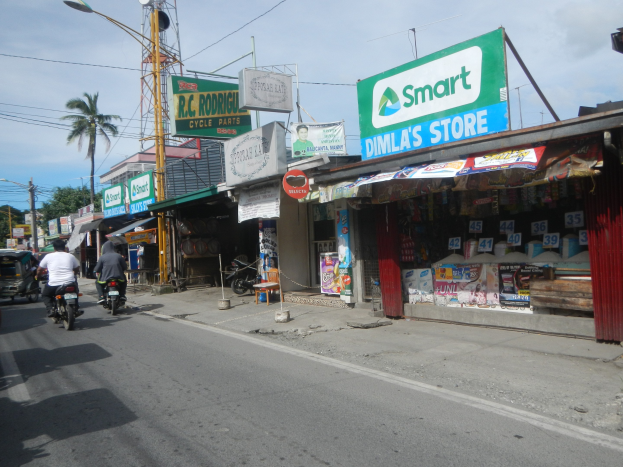 Eine belebte Straße mit Motorrädern, Autos, Gebäuden, Schildern, Strommasten, Drähte, Straßenlaternen, Bäume und einem bewölkten Himmel.
