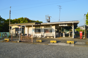 Ein Bahnhof mit Fahrrädern davor, eine Straße darunter und Häuser, Bäume, Pfähle, Drähte und einen klaren blauen Himmel im Hintergrund.