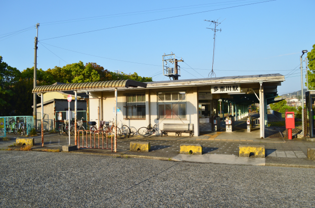 Ein Bahnhof mit Fahrrädern davor, eine Straße darunter und Häuser, Bäume, Pfähle, Drähte und einen klaren blauen Himmel im Hintergrund.