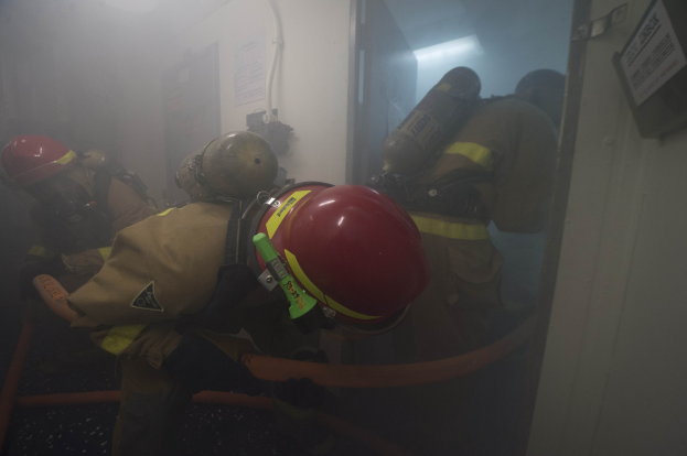 Feuerwehrleute in Schutzausrüstung mit Sauerstofftanks betreten einen raucherfüllten Raum, mit Rohren im Vorder- und Hintergrund.