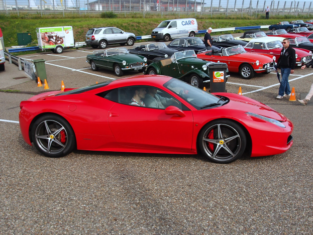 Eine Gruppe von Autos auf einem Parkplatz mit Menschen in der Nähe, umgeben von Verkehrskegeln, Mülltonnen und Schildern, mit einem Zaun, Grünfläche, Gebäuden und einem klaren Himmel im Hintergrund.