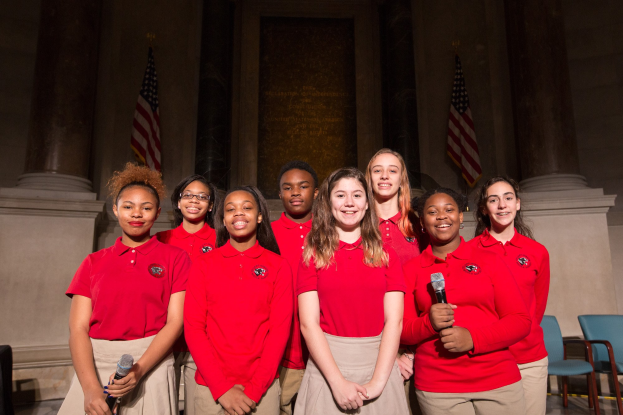 Eine Gruppe junger Frauen in roten Shirts, die strahlend Mikrofone halten und vor Stühlen, Säulen und Flaggen stehen, wahrscheinlich Teil eines nationalen Jugendchors.