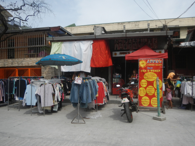 Eine Straßenszene mit einem Motorrad vor einem Bekleidungsgeschäft mit Kleiderbügeln, einem Schirm und einer Person in der Nähe, mit Gebäuden, einem Baum und Himmel im Hintergrund.