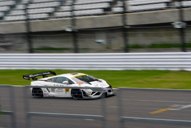 Weißer Sportwagen fährt mit hoher Geschwindigkeit auf einer Rennstrecke, umgeben von einem Zaun und Gras, mit einem unscharfen Hintergrund.