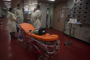 Gruppe von Menschen in Schutzausrüstung um eine Trage in einem Krankenhauszimmer mit medizinischer Ausrüstung, einem Bett, einem Ständer, einer Tür, einer Säule, einem Schild und Deckenleuchten.