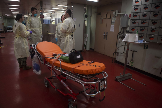 Gruppe von Menschen in Schutzausrüstung um eine Trage in einem Krankenhauszimmer mit medizinischer Ausrüstung, einem Bett, einem Ständer, einer Tür, einer Säule, einem Schild und Deckenleuchten.