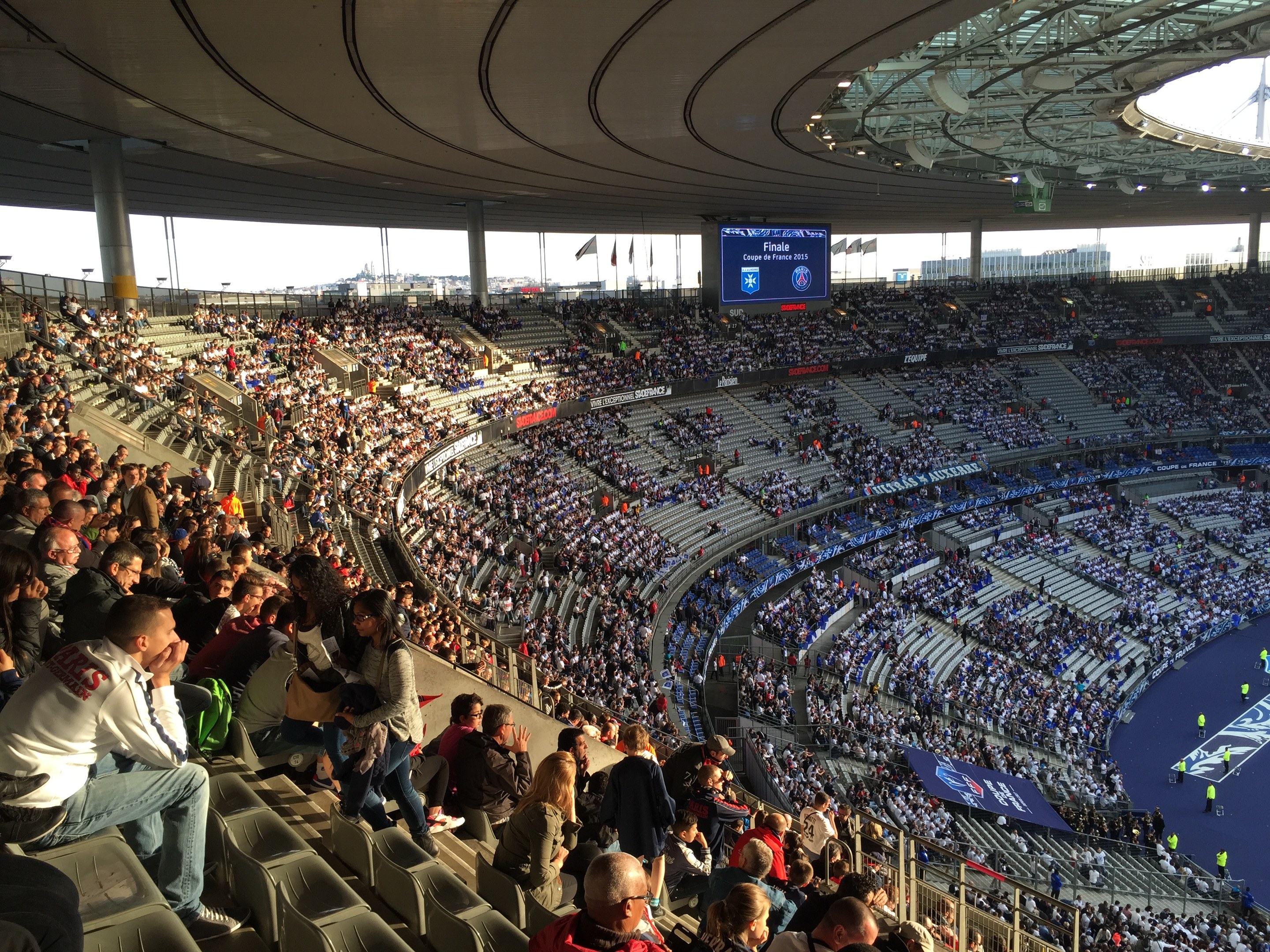Große Menschenmenge in einem Stadion bei einem Fußballspiel mit einer Bühne rechts, Fahnen, Stangen, einem Bildschirm und der Allianz Arena in München, Deutschland im Hintergrund.