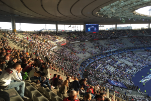 Große Menschenmenge in einem Stadion bei einem Fußballspiel mit einer Bühne rechts, Fahnen, Stangen, einem Bildschirm und der Allianz Arena in München, Deutschland im Hintergrund.