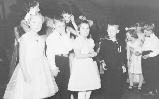 Schwarz-weißes Bild einer Gruppe von Kindern, die auf einer Tanzfläche stehen, lächeln und glücklich aussehen, mit einer Wand im Hintergrund.