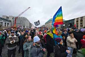 Eine große Gruppe von Menschen bei einer LGBTQ+-Rechtsdemo in Berlin, die Fahnen und Banner schwenken, mit Gebäuden und einem Kran im Hintergrund unter einem bewölkten Himmel.