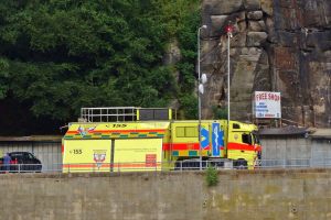 Rettungswagen auf der Seite einer Straße neben einer Klippe geparkt, mit verschiedenen Gegenständen wie einer Wand, einem Geländer, Pfosten, Lampen, Tafeln mit Text im Vordergrund und Bäumen und einem Auto im Hintergrund.