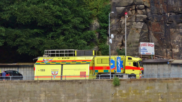 Rettungswagen auf der Seite einer Straße neben einer Klippe geparkt, mit verschiedenen Gegenständen wie einer Wand, einem Geländer, Pfosten, Lampen, Tafeln mit Text im Vordergrund und Bäumen und einem Auto im Hintergrund.