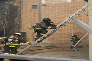 Feuerwehrleute in Helmen und Ausrüstung klettern an einer Leiter vor einem Backsteingebäude mit Rohren auf dem Boden und einer Metallstange an der Basis, während im Hintergrund ein weiteres Gebäude mit Fenstern und Netz zu sehen ist.