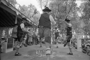 Eine Gruppe von Männern in traditioneller bayrischer Tracht tanzt auf einer Bühne vor einem gemischten Publikum, mit Bäumen und einem Schuppen im Hintergrund, dargestellt in Schwarz-Weiß.