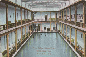 Schwarz-weiß-Postkarte eines prunkvollen Indoor-Schwimmbads im West Baden Springs Hotel mit Schwimmern, Säulen, Geländern, Stühlen und Text am unteren Rand.
